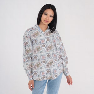 May include: A light blue button-down shirt with a repeating pattern of compasses, maps, and nautical elements in red, green, and brown. The long-sleeved shirt is made of a lightweight fabric.