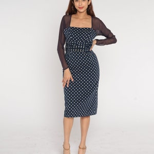 May include: A navy blue dress with white polka dots and a sheer black overlay. The dress has a square neckline and a fitted silhouette. The model is wearing brown heeled sandals.