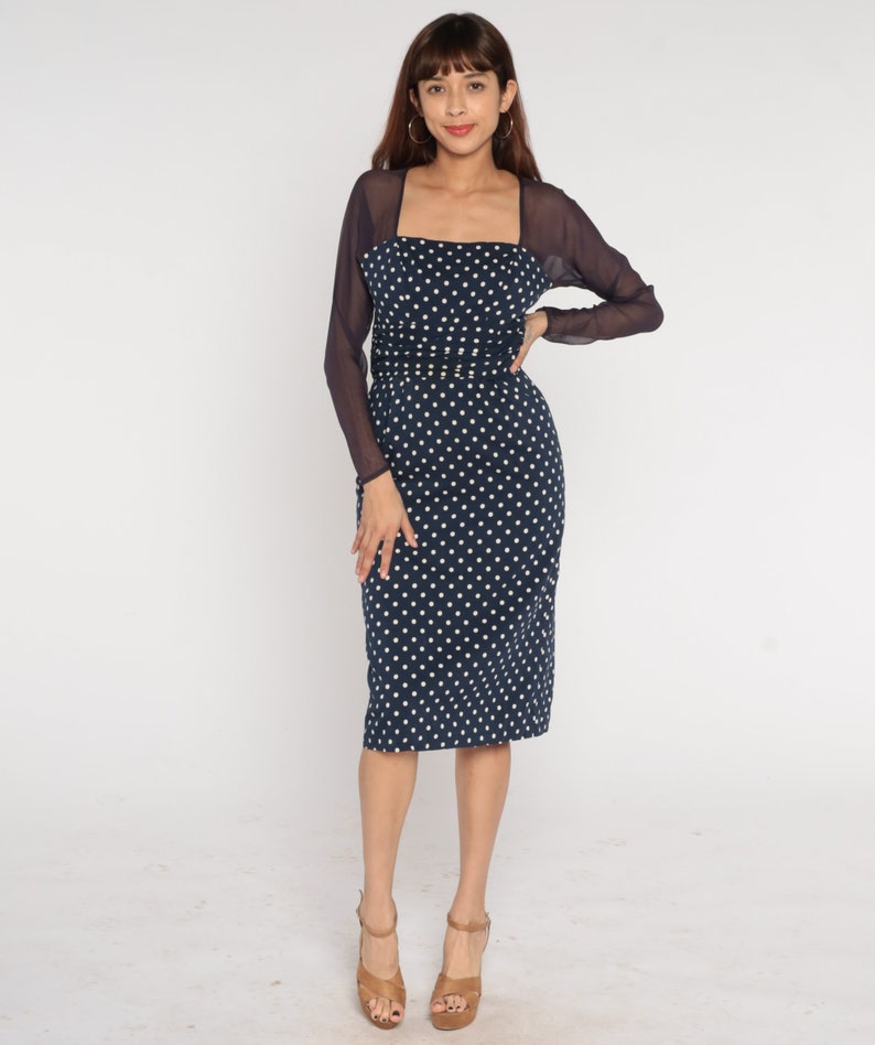 May include: A navy blue dress with white polka dots and a sheer black overlay. The dress has a square neckline and a fitted silhouette. The model is wearing brown heeled sandals.