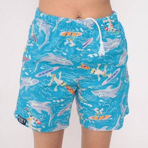 K&ouml;nnte beinhalten: Blaue Badehose mit wei&szlig;em Kordelzug und einem bunten Druck von Haien, Surfern und Wellen. Der Text "KIWI SURF" ist auf dem linken Bein aufgedruckt.