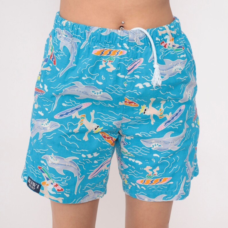 K&ouml;nnte beinhalten: Blaue Badehose mit wei&szlig;em Kordelzug und einem bunten Druck von Haien, Surfern und Wellen. Der Text "KIWI SURF" ist auf dem linken Bein aufgedruckt.