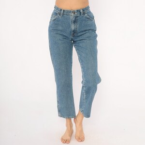 May include: Light blue denim jeans with a classic straight leg fit. The jeans have a button and zipper closure, front and back pockets, and belt loops. The jeans are a medium wash and appear to be vintage.
