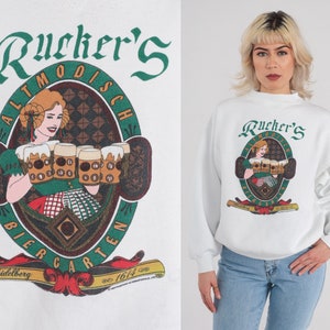 May include: White sweatshirt with a green and red graphic of a woman holding beer steins. The graphic says "Rucker's Altmodisch Biergarten Heidelberg est. 1614".