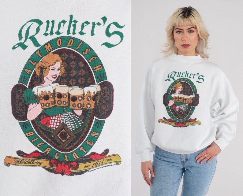 May include: White sweatshirt with a green and red graphic of a woman holding beer steins. The graphic says "Rucker's Altmodisch Biergarten Heidelberg est. 1614".