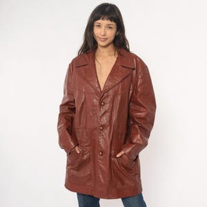 May include: A reddish-brown leather jacket with a collared neckline, three buttons, and two front pockets. The jacket is knee-length and has a vintage style.