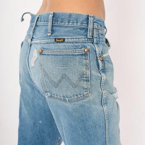 May include: Light blue denim Wrangler jeans with distressed details. The jeans feature a classic five-pocket design, copper rivets, and the Wrangler logo on the back pocket. The jeans have a faded wash and a worn appearance.