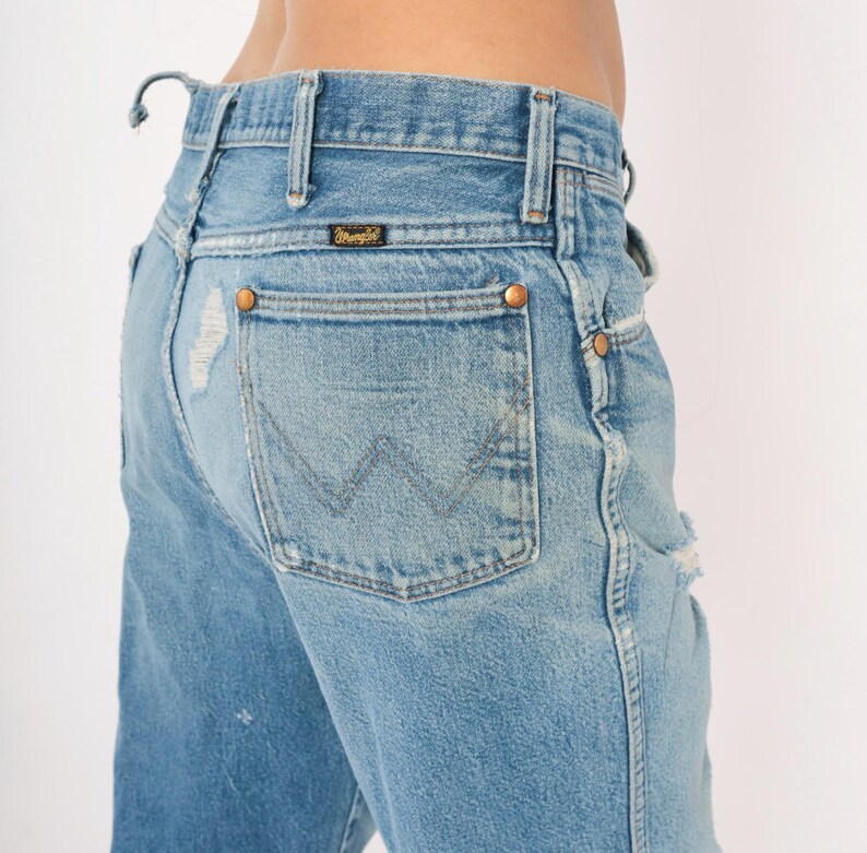 May include: Light blue denim Wrangler jeans with distressed details. The jeans feature a classic five-pocket design, copper rivets, and the Wrangler logo on the back pocket. The jeans have a faded wash and a worn appearance.