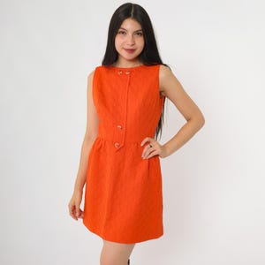 May include: A sleeveless orange dress with a quilted pattern. The dress features a round neckline, gold-colored buttons, and a fitted waist. The dress is knee-length and has a simple, classic design.