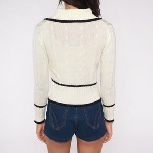 May include: Cream-colored sweater with black trim. The sweater has a high collar and long sleeves. The black trim is on the collar, waist, and cuffs. The sweater is paired with blue denim shorts.