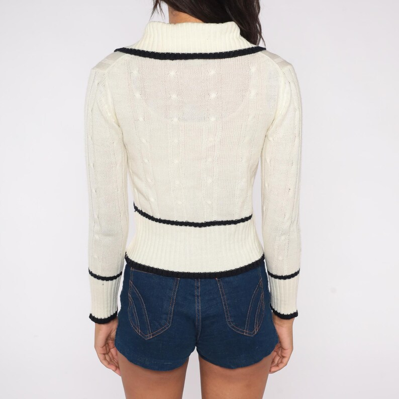 May include: Cream-colored sweater with black trim. The sweater has a high collar and long sleeves. The black trim is on the collar, waist, and cuffs. The sweater is paired with blue denim shorts.