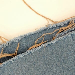May include: Close-up of light blue denim fabric with frayed edges and tan twine accents. The denim has a textured appearance, and the twine adds a rustic touch. The image suggests a craft or textile project.