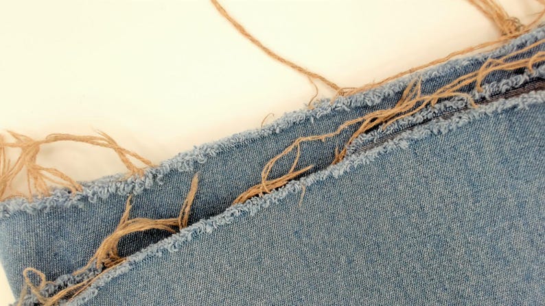 May include: Close-up of light blue denim fabric with frayed edges and tan twine accents. The denim has a textured appearance, and the twine adds a rustic touch. The image suggests a craft or textile project.