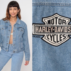 May include: Light blue denim jacket with a classic collar, button-down front, and chest pockets. The back of the jacket features a Harley-Davidson Cycles patch in black, white, and gray. The jacket is part of a denim clothing set.