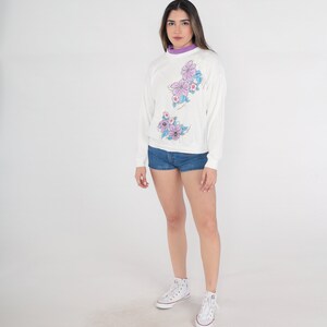 May include: A white sweatshirt with a floral design in shades of purple and blue. The sweatshirt has long sleeves and a crew neck. The model is wearing denim shorts and white trainers.