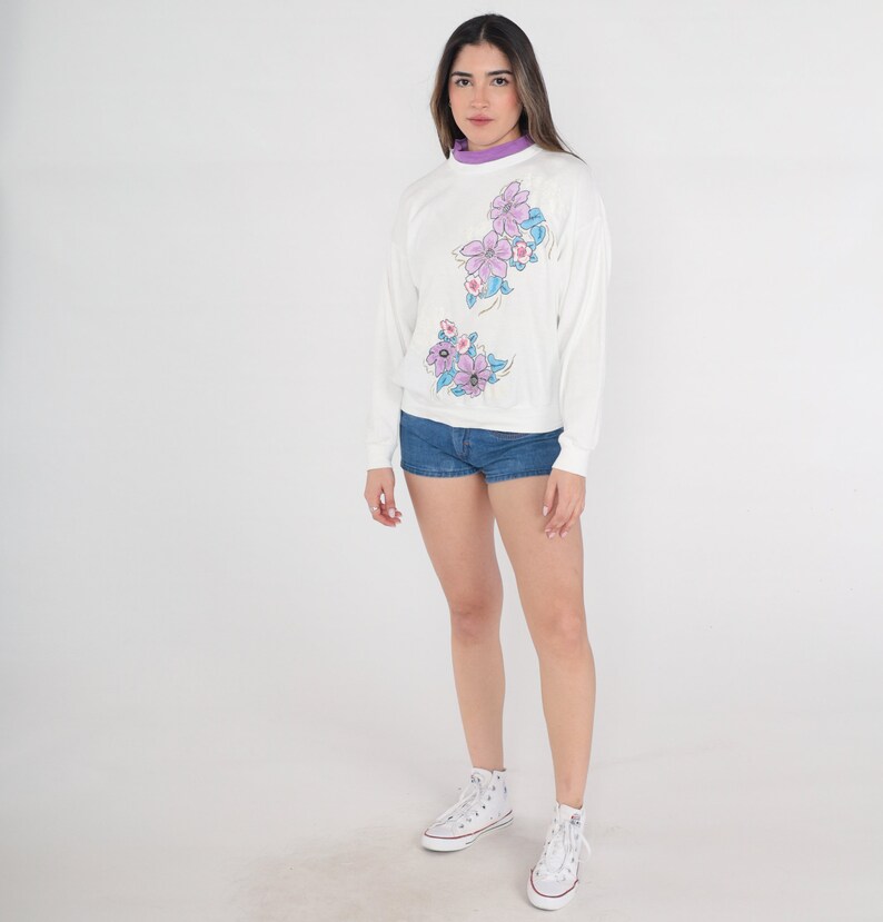 May include: A white sweatshirt with a floral design in shades of purple and blue. The sweatshirt has long sleeves and a crew neck. The model is wearing denim shorts and white trainers.