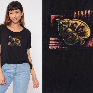 May include: Black short-sleeved t-shirt with a colorful leaf design on the front. The leaf is outlined in black and has shades of yellow, green, and pink.