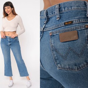 May include: A pair of light blue denim jeans with a brown leather patch that says "Wrangler" on the back pocket. The jeans have a straight leg and a frayed hem.