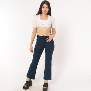 May include: A white cropped blouse with a scoop neckline, short sleeves featuring lace trim, and a front button closure, paired with dark blue flared trousers. The trousers have a high-waisted fit and a solid color finish, complemented by black platform shoes.