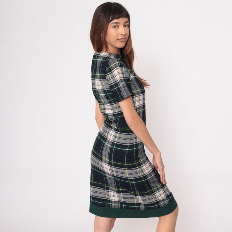May include: A plaid dress in a green, navy, and cream pattern. The dress has short sleeves and a knee-length hem with a dark green border. The fabric has a classic tartan design, suitable for casual wear.