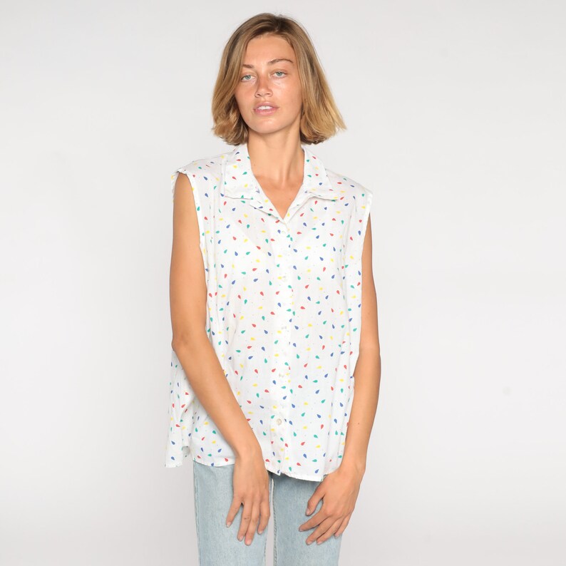 May include: White sleeveless button-down shirt with a colorful leaf pattern. The shirt features a collared neckline and a relaxed fit. The pattern includes red, yellow, blue, and green leaf shapes.