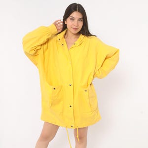 May include: A bright yellow jacket featuring a loose, oversized fit, long sleeves with cuffed wrists, a front button-down closure with metallic snaps, a pointed collar, and two large patch pockets on the front, finished with a drawstring hem.