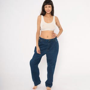 May include: A woman wearing a cream-colored tank top and dark blue denim jeans. The jeans have a high waist and a relaxed fit. The background is plain white.