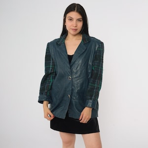 May include: Teal leather blazer with plaid sleeves. The blazer has a notched collar, two gold buttons, and a tailored fit. The sleeves are a dark green, black, and red plaid pattern. The blazer is worn over a black dress.