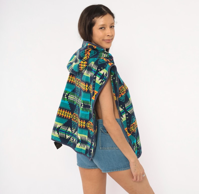 May include: A hooded poncho featuring a vibrant geometric pattern in shades of teal, navy blue, yellow, and orange. The garment has a dark trim along the edges and is worn over blue denim shorts. The fabric displays a repeating angular design.