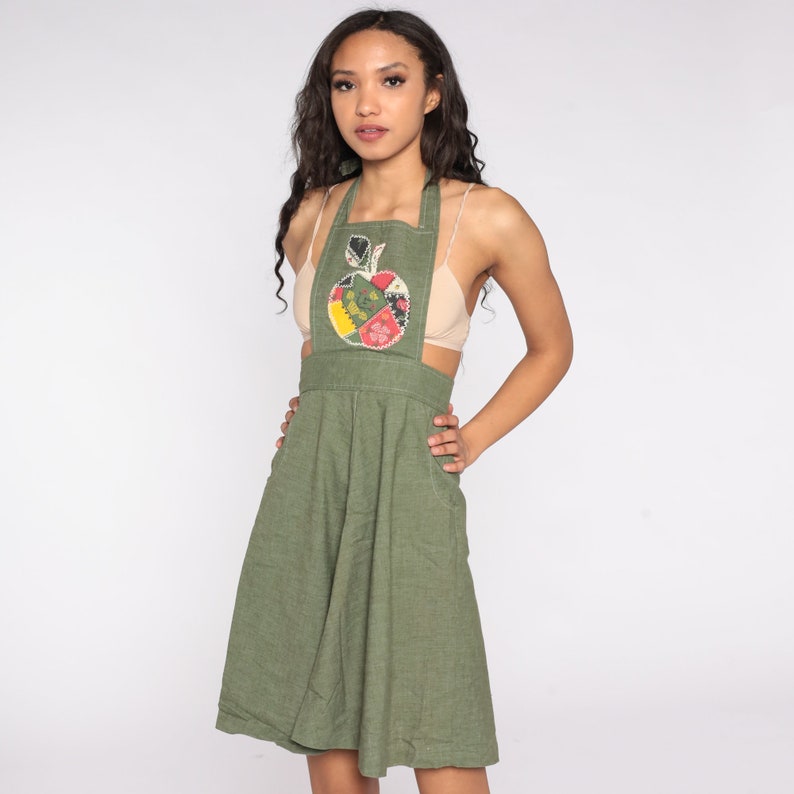 May include: Olive green apron dress with a colorful apple applique. The dress features a bib top, a fitted waist, and a flared skirt. It has pockets on the sides and adjustable straps. The apple applique has red, yellow, and green patches.