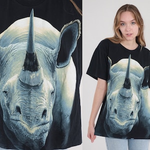 以下が含まれることがあります： 大きなサイの顔のグラフィックプリントが施された黒いTシャツ。サイは大きな角を持ち、灰色と白の色合いで描かれています。