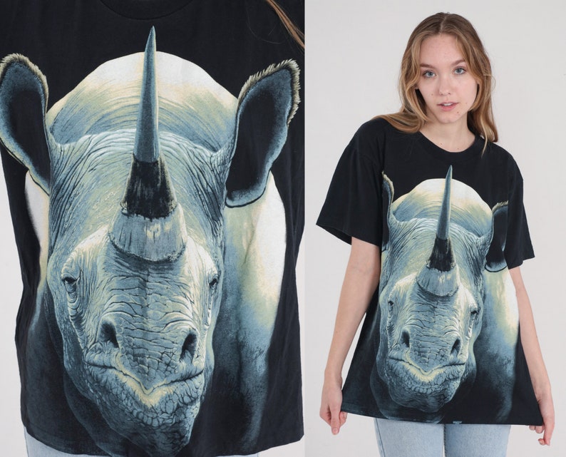 以下が含まれることがあります： 大きなサイの顔のグラフィックプリントが施された黒いTシャツ。サイは大きな角を持ち、灰色と白の色合いで描かれています。