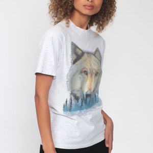 May include: A gray t-shirt with a wolf graphic print. The wolf is looking directly at the viewer and is surrounded by a blue and green forest scene.