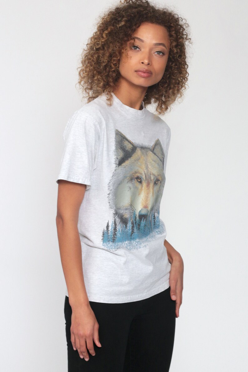 May include: A gray t-shirt with a wolf graphic print. The wolf is looking directly at the viewer and is surrounded by a blue and green forest scene.