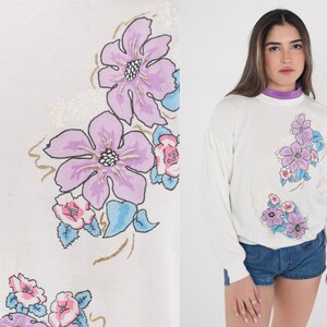 May include: White long-sleeved sweatshirt with a floral print featuring pink, purple and blue flowers. The flowers are stylised and have a vintage look.