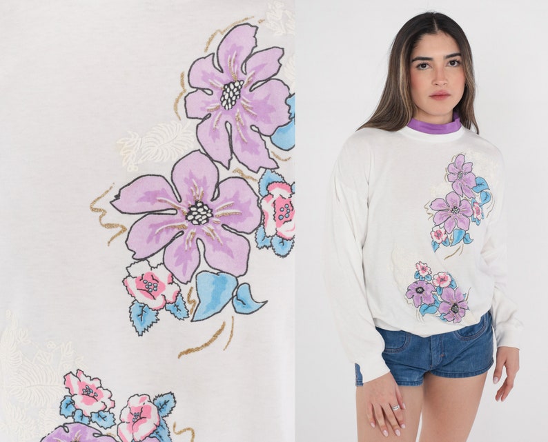 May include: White long-sleeved sweatshirt with a floral print featuring pink, purple and blue flowers. The flowers are stylised and have a vintage look.