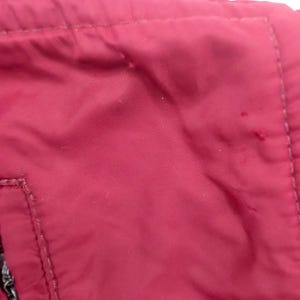 May include: Close-up of a section of a maroon jacket. The fabric has a slight sheen and is stitched together with matching thread. A small pocket with a zipper is visible, along with a seam.