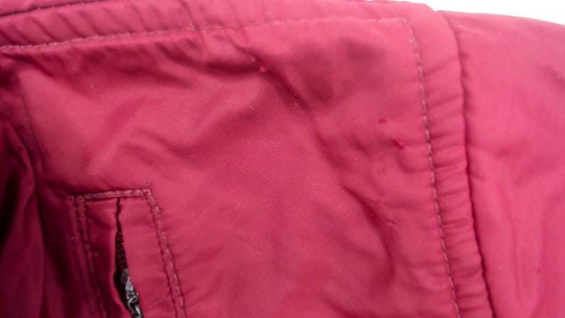 May include: Close-up of a section of a maroon jacket. The fabric has a slight sheen and is stitched together with matching thread. A small pocket with a zipper is visible, along with a seam.