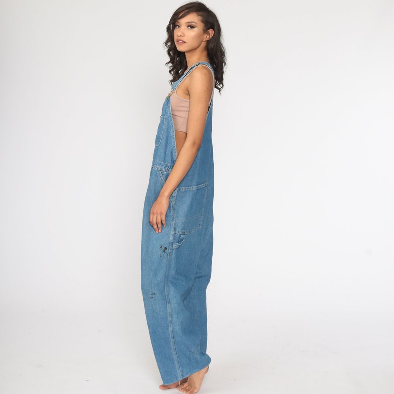 May include: A woman wearing a light blue denim overall with a light brown tank top underneath. The overall has a wide leg and is slightly distressed.
