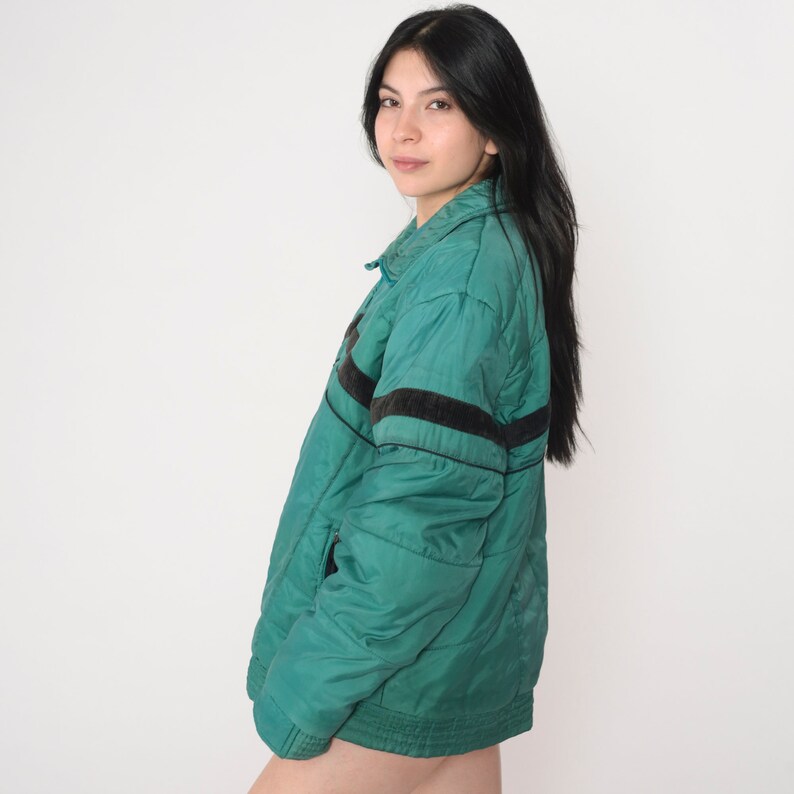 May include: A teal blue quilted jacket with black and white stripes on the sleeves. The jacket has a zipper closure and a stand-up collar.