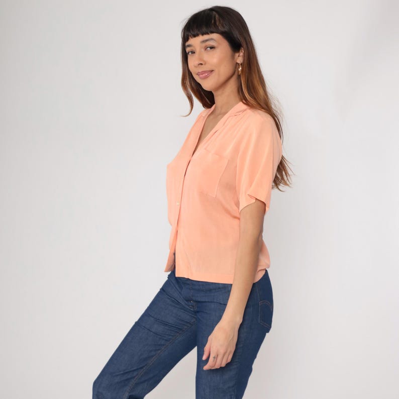Puede incluir: Una camisa melocot&oacute;n de manga corta con cuello y un bolsillo en el pecho. La camisa est&aacute; abotonada por delante y se combina con unos vaqueros azules. La modelo est&aacute; de pie frente a un fondo blanco.
