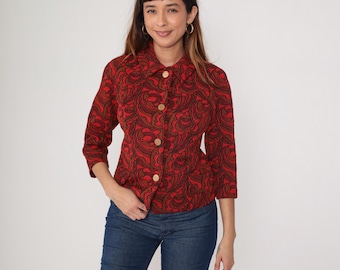 May include: A red and brown patterned button-up jacket with three-quarter sleeves. The jacket has a paisley-like design and is buttoned up with gold-colored buttons. The person is wearing blue jeans.