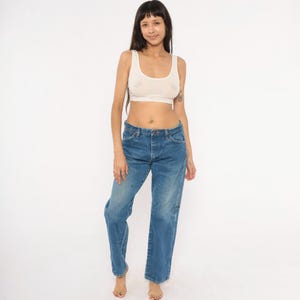 Pode incluir: Uma mulher vestindo uma regata branca e jeans azul. Os jeans t&ecirc;m fecho de bot&atilde;o e corte reto. A regata &eacute; um design simples e sem mangas.