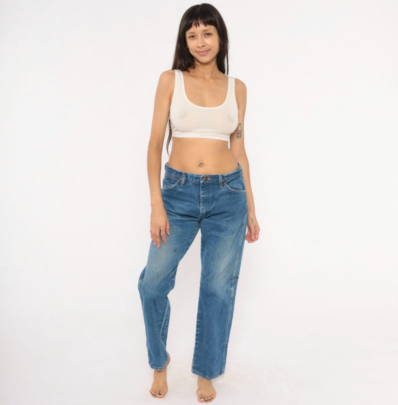 Pode incluir: Uma mulher vestindo uma regata branca e jeans azul. Os jeans t&ecirc;m fecho de bot&atilde;o e corte reto. A regata &eacute; um design simples e sem mangas.