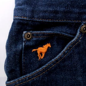 May include: Close-up of dark blue denim jeans pocket with an orange embroidered horse logo. The pocket has a metal button and is stitched with a contrasting thread. The jeans are made of a durable denim fabric.