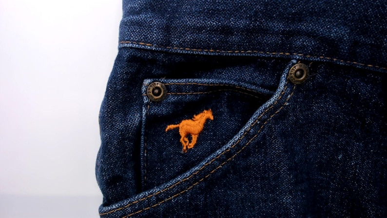 May include: Close-up of dark blue denim jeans pocket with an orange embroidered horse logo. The pocket has a metal button and is stitched with a contrasting thread. The jeans are made of a durable denim fabric.