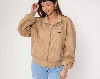 May include: Tan bomber jacket with a zip-up front, tan collar, and tan ribbed cuffs and hem. The jacket has a small pocket on the chest and a logo patch. The model is wearing light blue jeans.