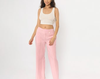 Puede incluir: Una mujer con una camiseta sin mangas blanca y pantalones rosas de pierna ancha. Los pantalones tienen cintura alta y dos botones. La mujer lleva sandalias de plataforma marrones. El fondo es blanco.