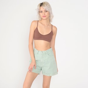 Puede incluir: Una mujer con un top de tirantes marrón de canalé y pantalones cortos de mezclilla verde claro. Está de pie frente a un fondo blanco.