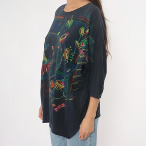 May include: Dark navy blue long-sleeve shirt with a colorful abstract design. The artwork features shapes in red, green, yellow, and blue. The shirt has a round neckline and three-quarter sleeves.