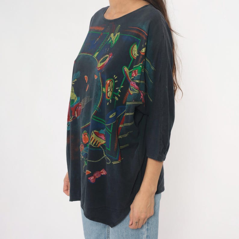 May include: Dark navy blue long-sleeve shirt with a colorful abstract design. The artwork features shapes in red, green, yellow, and blue. The shirt has a round neckline and three-quarter sleeves.